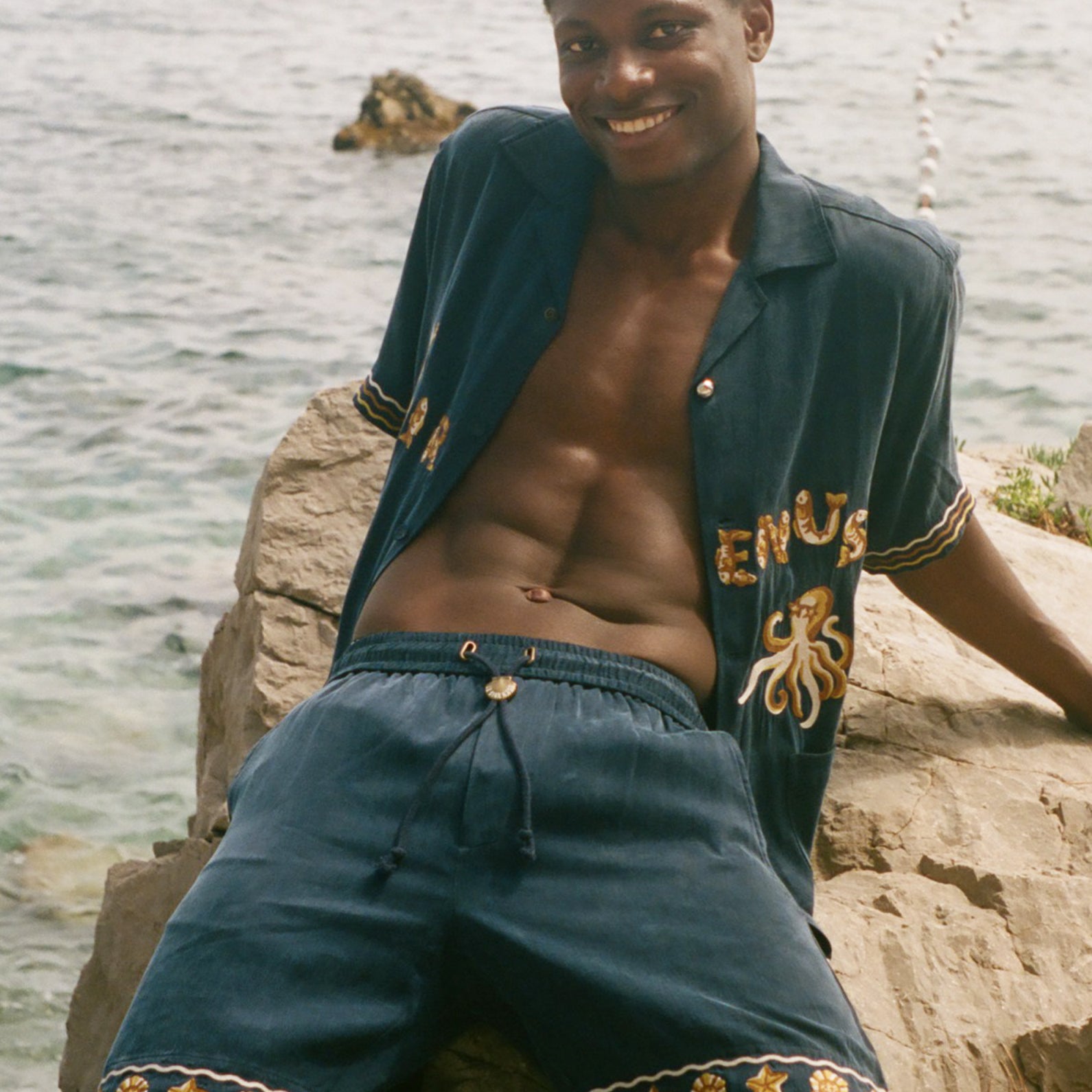Man wearing a blue shirt and shorts sitting on rocks by the water