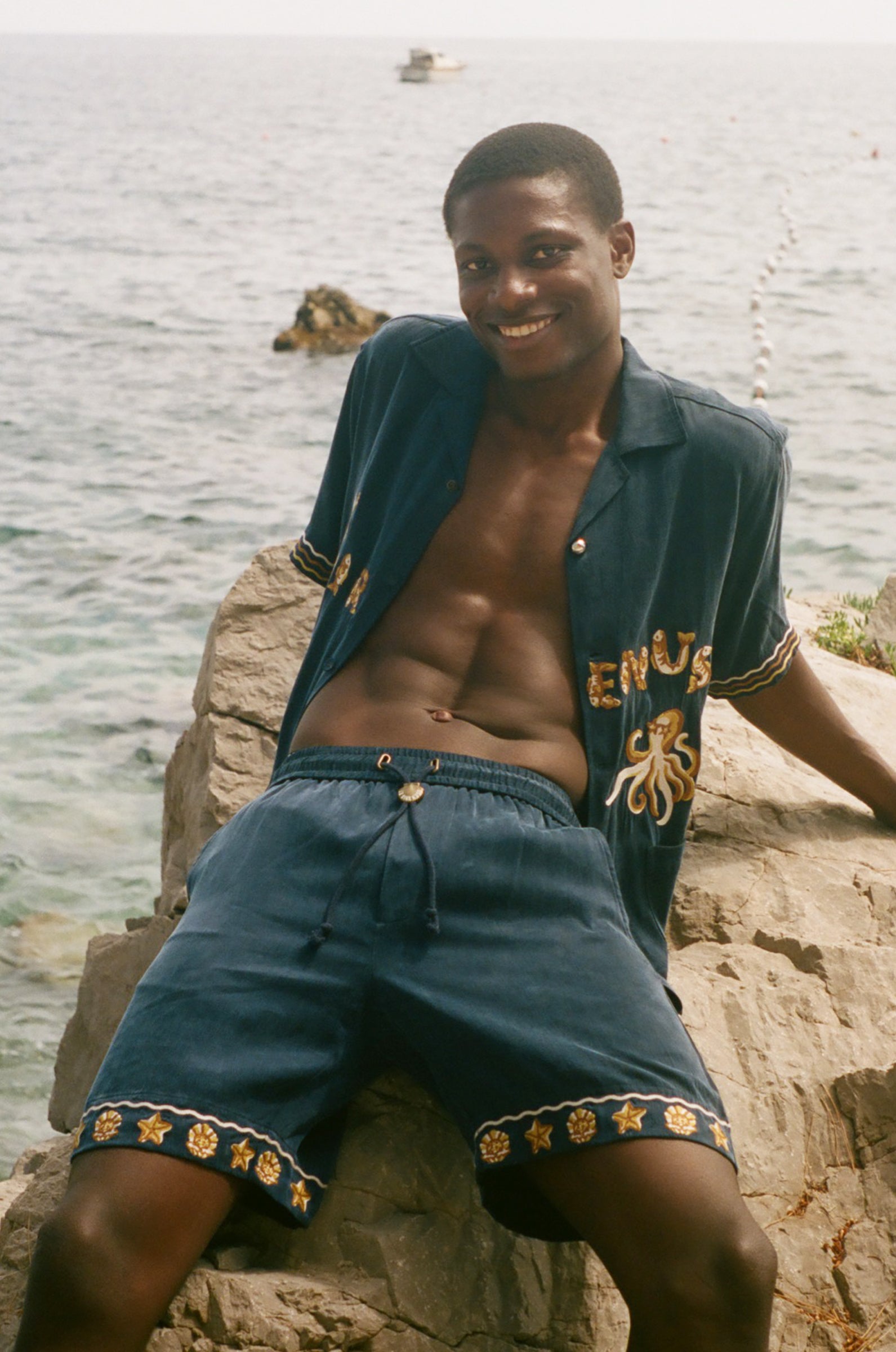 Man wearing a blue shirt and shorts sitting on rocks by the water