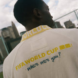 Person wearing a beige shirt with 'FIFA World Cup 2026' text on the back, standing outdoors with a cityscape in the background.