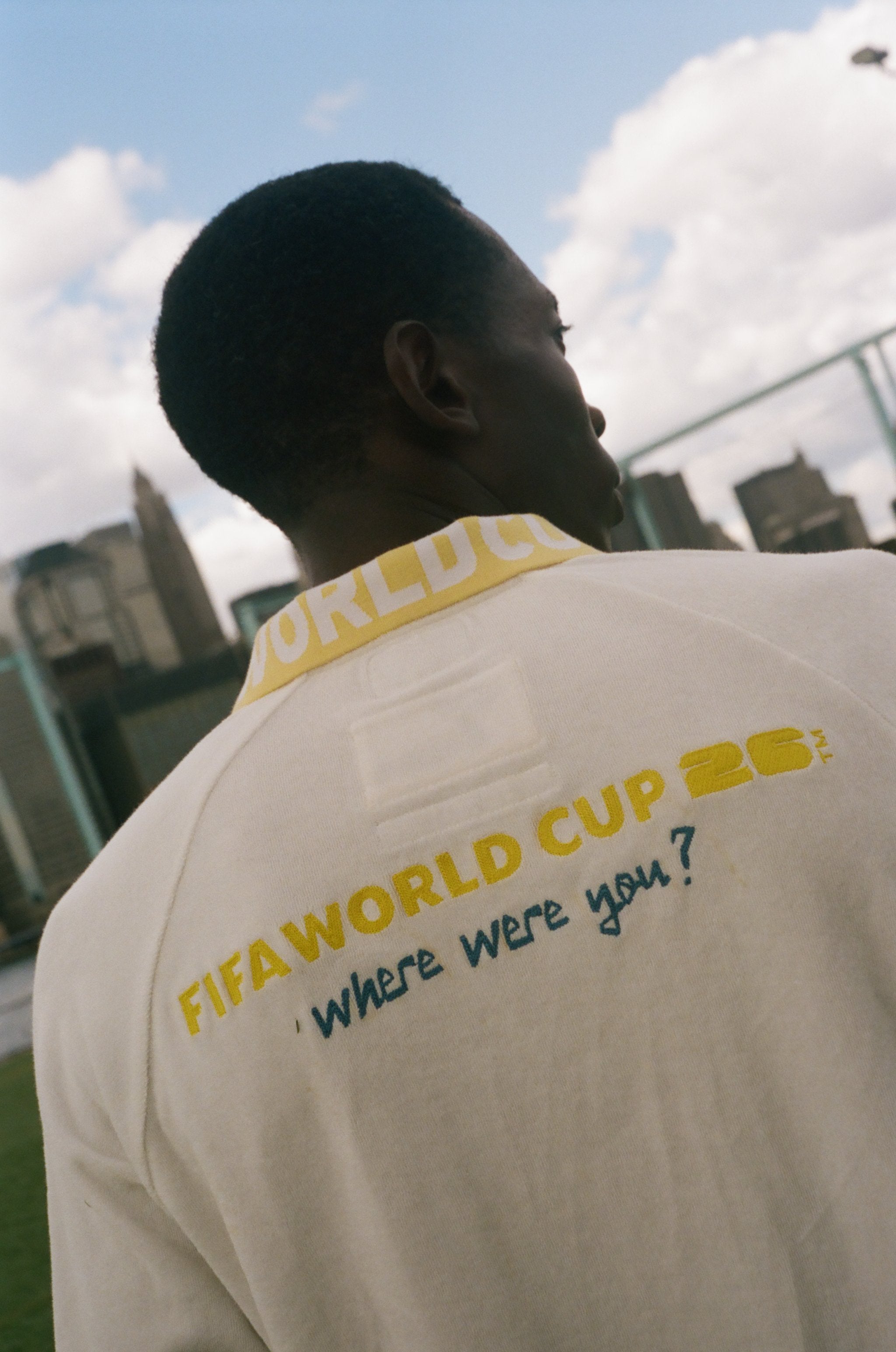 Person wearing a beige shirt with 'FIFA World Cup 2026' text on the back, standing outdoors with a cityscape in the background.