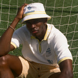Person wearing a white bucket hat and white polo shirt with yellow accents, sitting on grass.