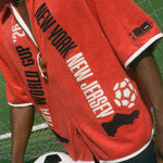 Person wearing a red 'New York New Jersey' World Cup Cabana holding a soccer ball on a grassy field.