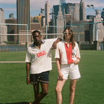 Two people standing on a grassy field with a city skyline in the background, wearing items from the FIFA and Tombolo collection.