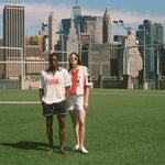 Two people standing on a grassy field with a city skyline in the background, wearing FIFA and Tombolo collection items. 