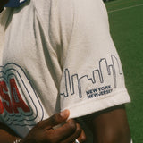 Person wearing a white terry cloth cabana shirt with 'USA' and skyline design on sleeve, standing on a grassy field.