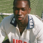 Person wearing a white cabana shirt with 'USA' and FIFA logo on a grass field.