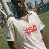 Man wearing a white cabana shirt with 'USA' logo on a sports field with city skyline in the background