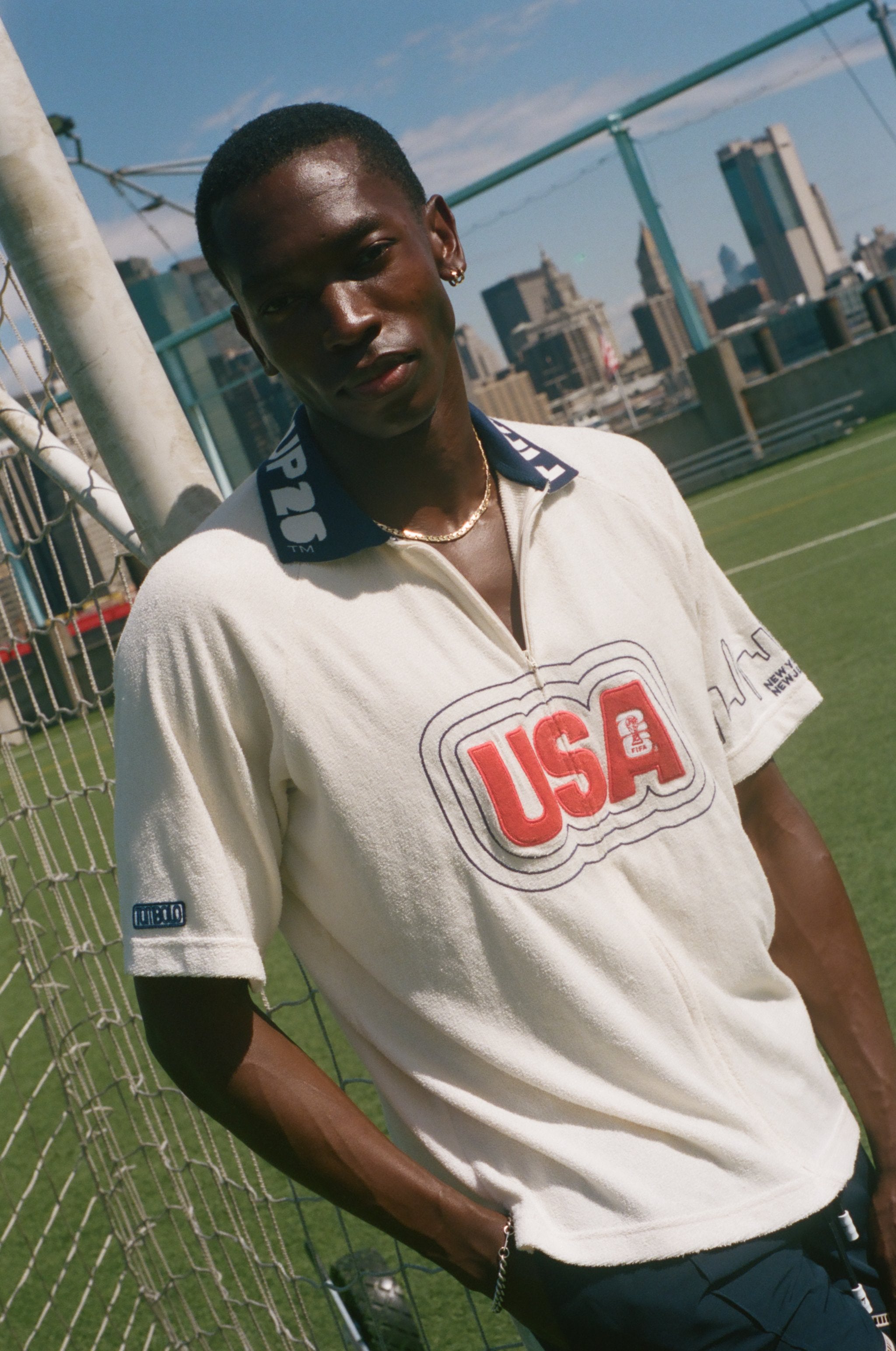 Man wearing a white cabana shirt with 'USA' logo on a sports field with city skyline in the background
