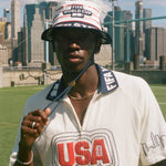 Person wearing a white shirt with 'USA' on it, holding the string to a USA bucket hat, with a city skyline in the background.