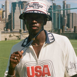 Person wearing a white shirt with 'USA' on it, holding the string to a USA bucket hat, with a city skyline in the background.