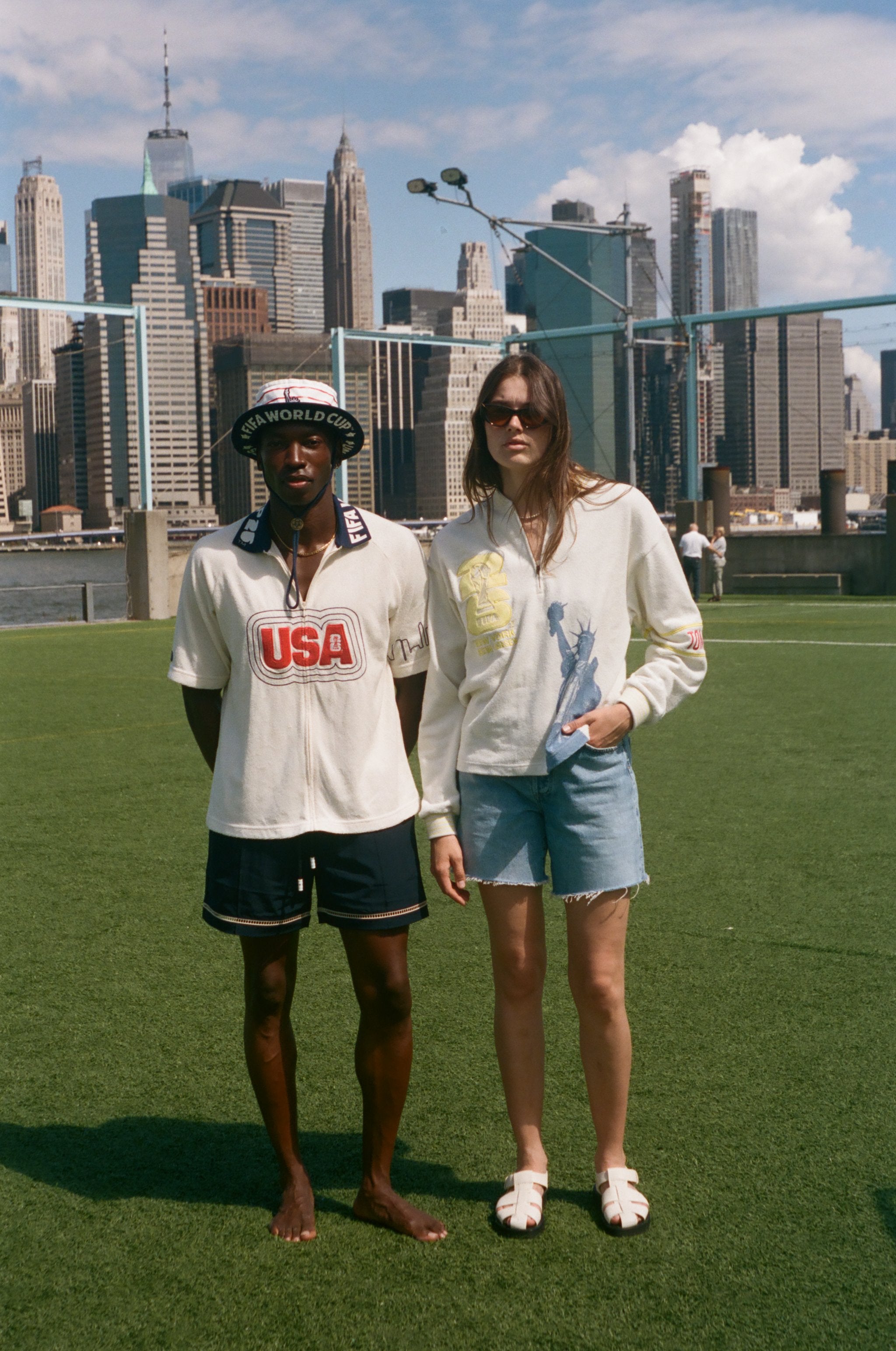 Two people standing on a grassy area with a city skyline in the background, wearing items from the FIFA and Tombolo collection.