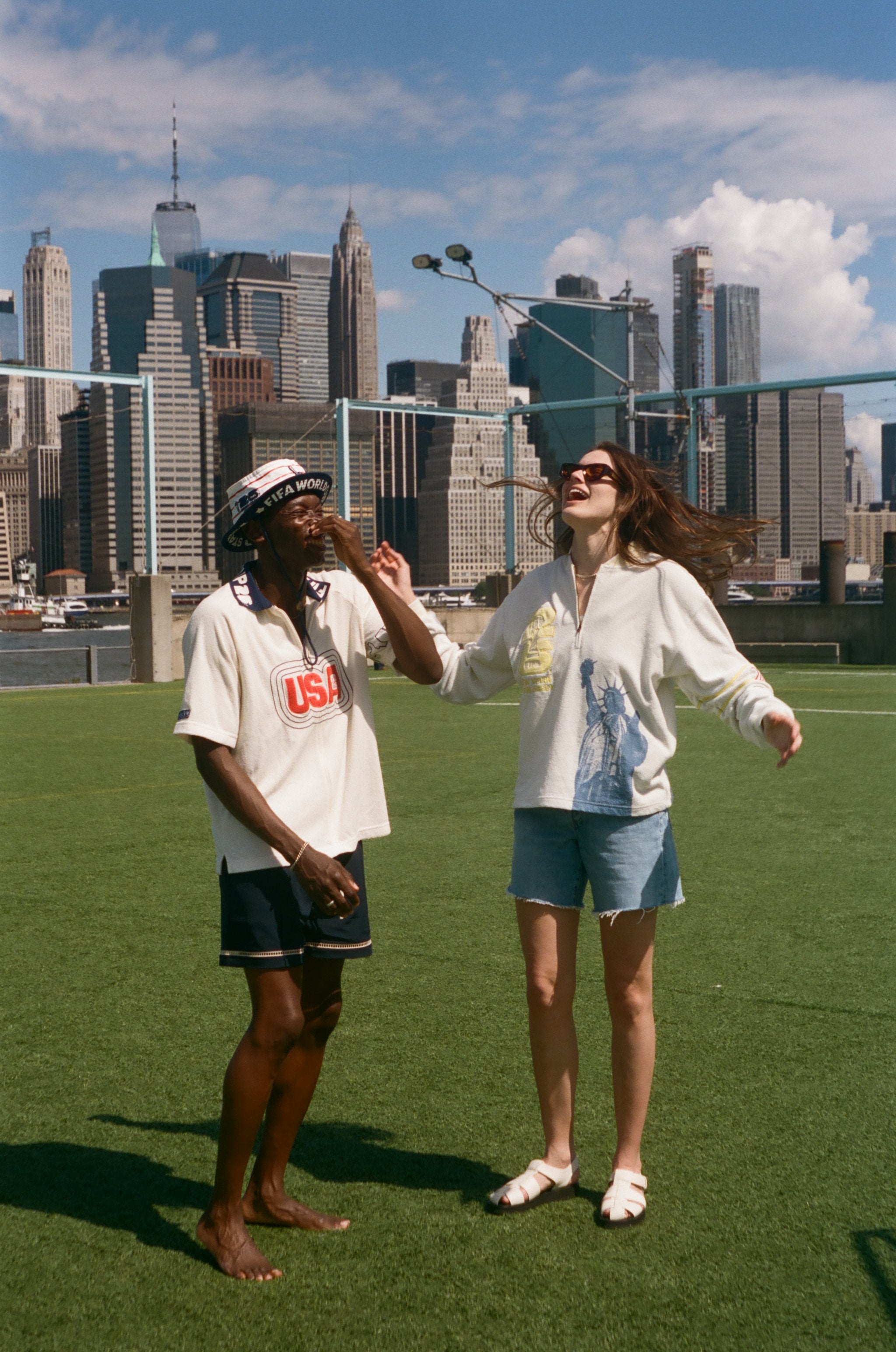 Two people standing on a grassy area with a city skyline in the background, wearing items from the FIFA and Tombolo collection.