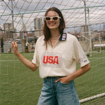 Person wearing a 'USA' cabana shirt and denim shorts standing in front of a sports net with a cityscape in the background.