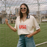 Person wearing a 'USA' cabana shirt and denim shorts standing in front of a sports net with a cityscape in the background.