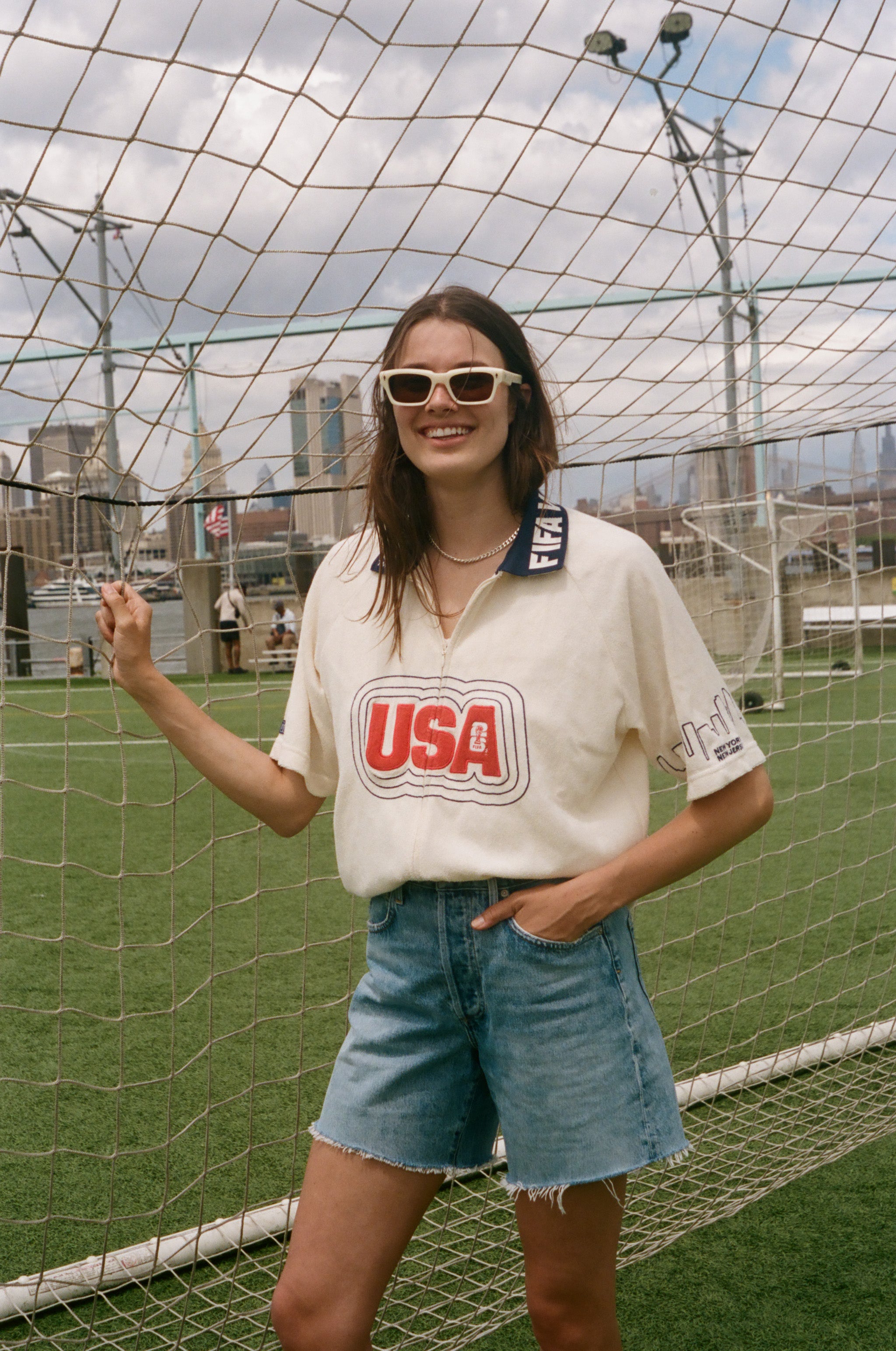 Person wearing a 'USA' cabana shirt and denim shorts standing in front of a sports net with a cityscape in the background.