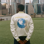 Person wearing a white jacket with a world cup design, standing on a grassy field with a city skyline in the background.