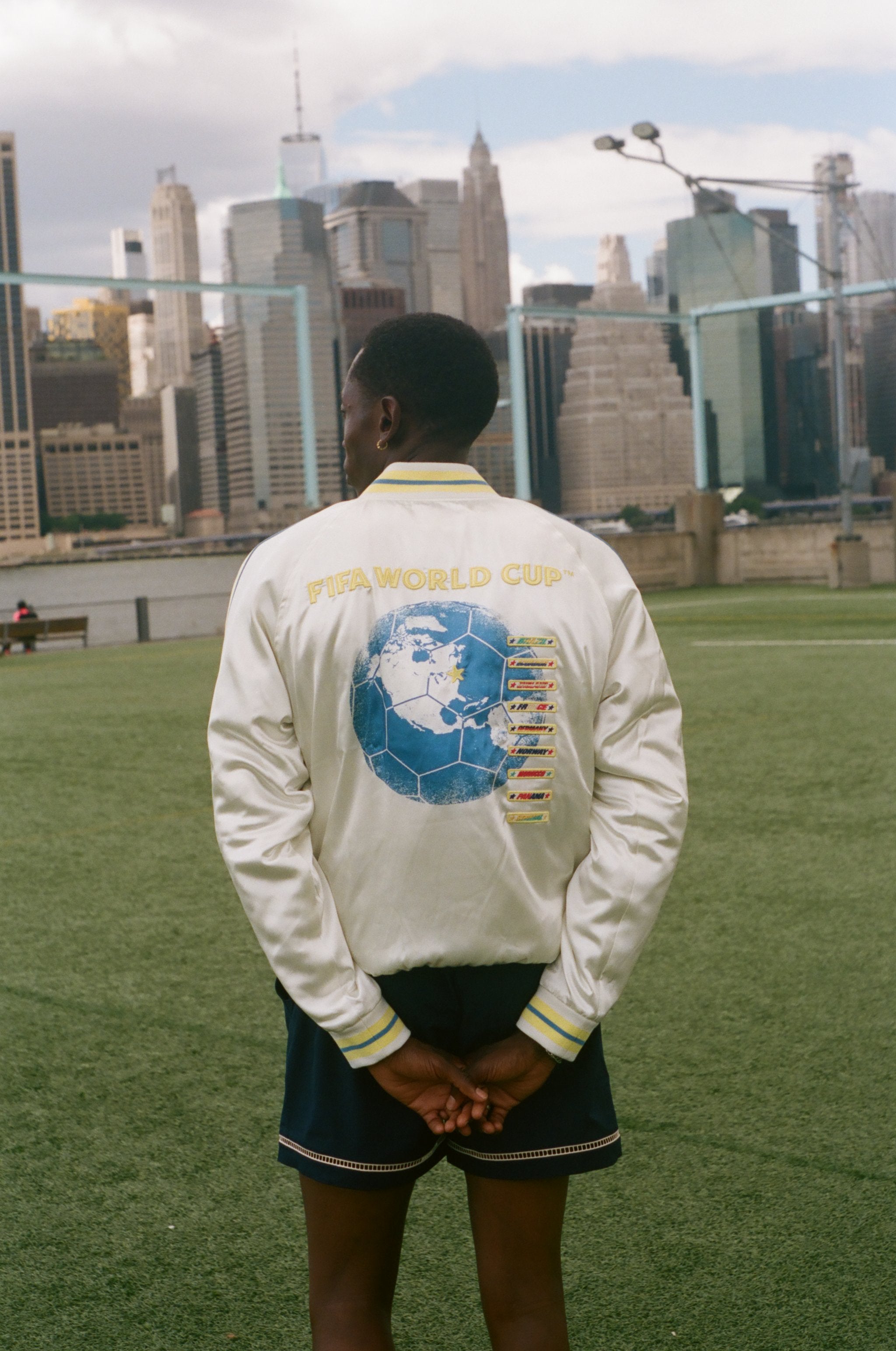 Person wearing a white jacket with a world cup design, standing on a grassy field with a city skyline in the background.