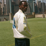 Person wearing a white jacket with a world cup design, standing on a grassy field with a city skyline in the background.