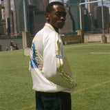 Person wearing a white jacket with a world cup design, standing on a grassy field with a city skyline in the background.