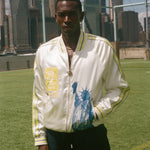 Person wearing a white jacket with a graphic statue of liberty on the front on a sports field with city skyline in the background