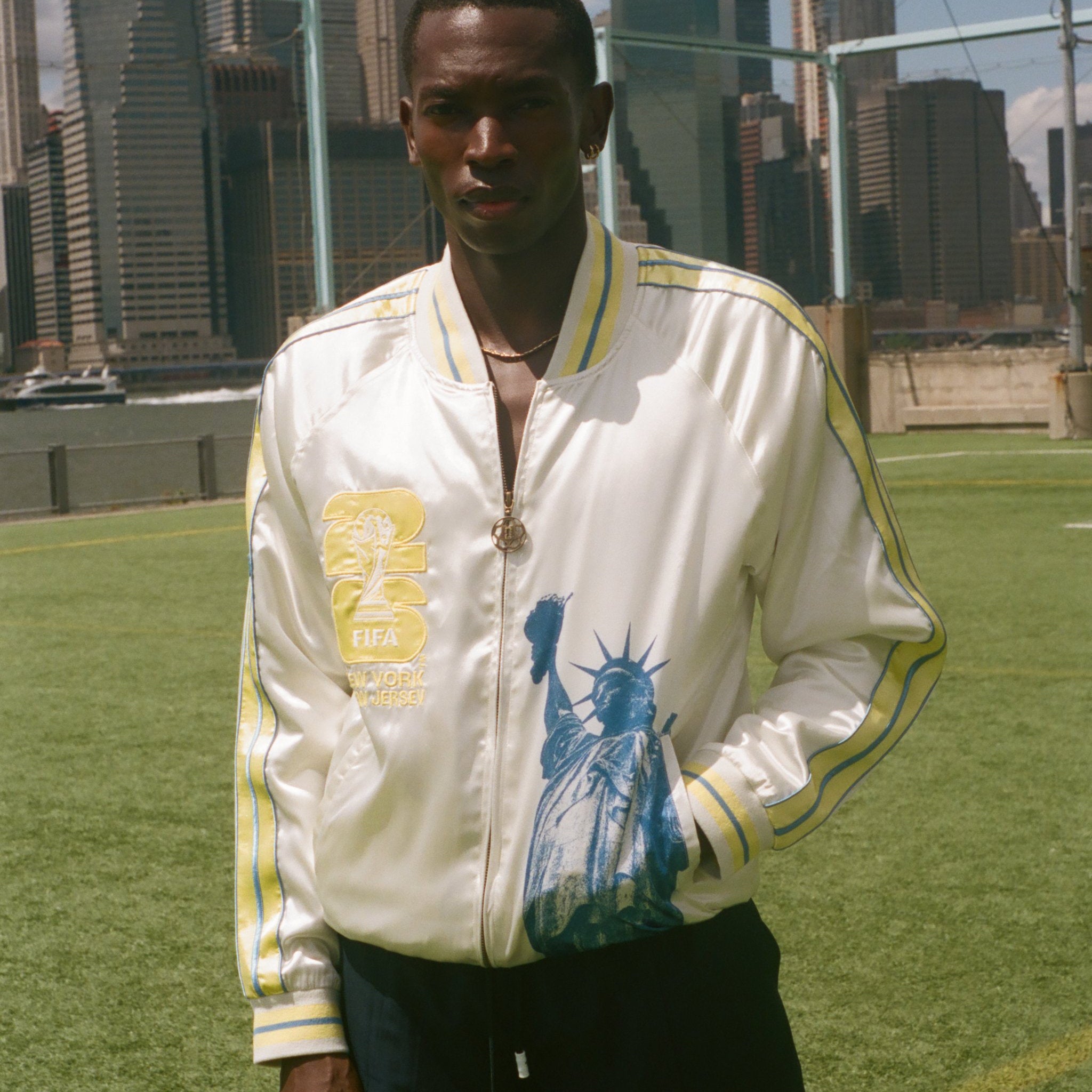 Person wearing a white jacket with a graphic statue of liberty on the front on a sports field with city skyline in the background