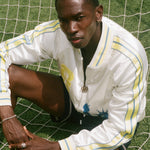 Person wearing a white FIFA souvenir jacket with yellow accents on a grassy field.