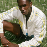 Person wearing a white FIFA souvenir jacket with yellow accents on a grassy field.