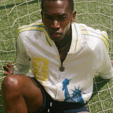 Man wearing FIFA Souvenir Jacket and swim trunks, sitting on a soccer net on a field. 