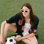 Woman sitting on grass with a soccer ball, wearing sunglasses and a navy USA FIFA cabana from the Tombolo and FIFA collection.
