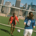 Two people playing soccer on a grassy field with a city skyline in the background, both wearing items from Tombolo and FIFA.