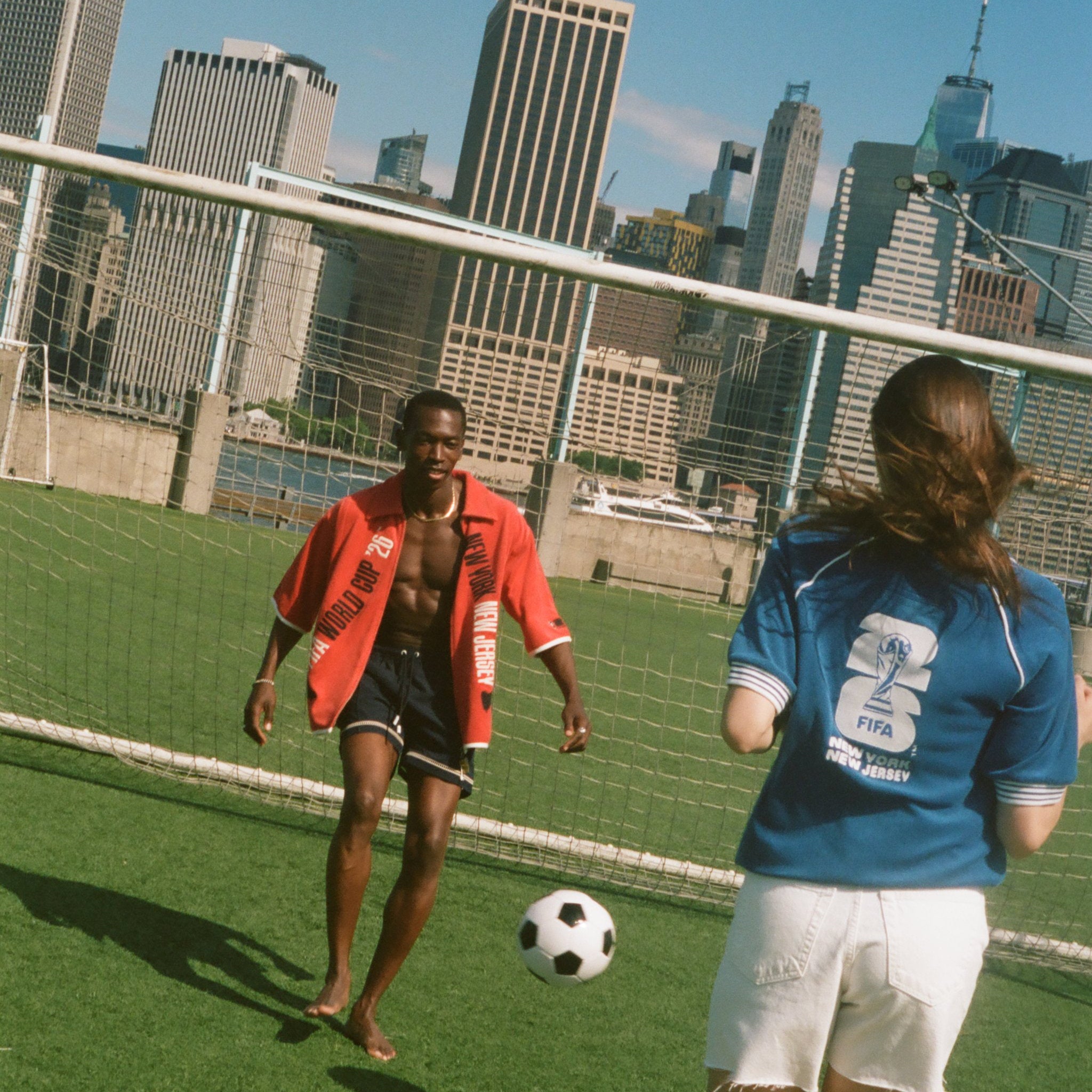 Two people playing soccer on a grassy field with a city skyline in the background, both wearing items from Tombolo and FIFA.