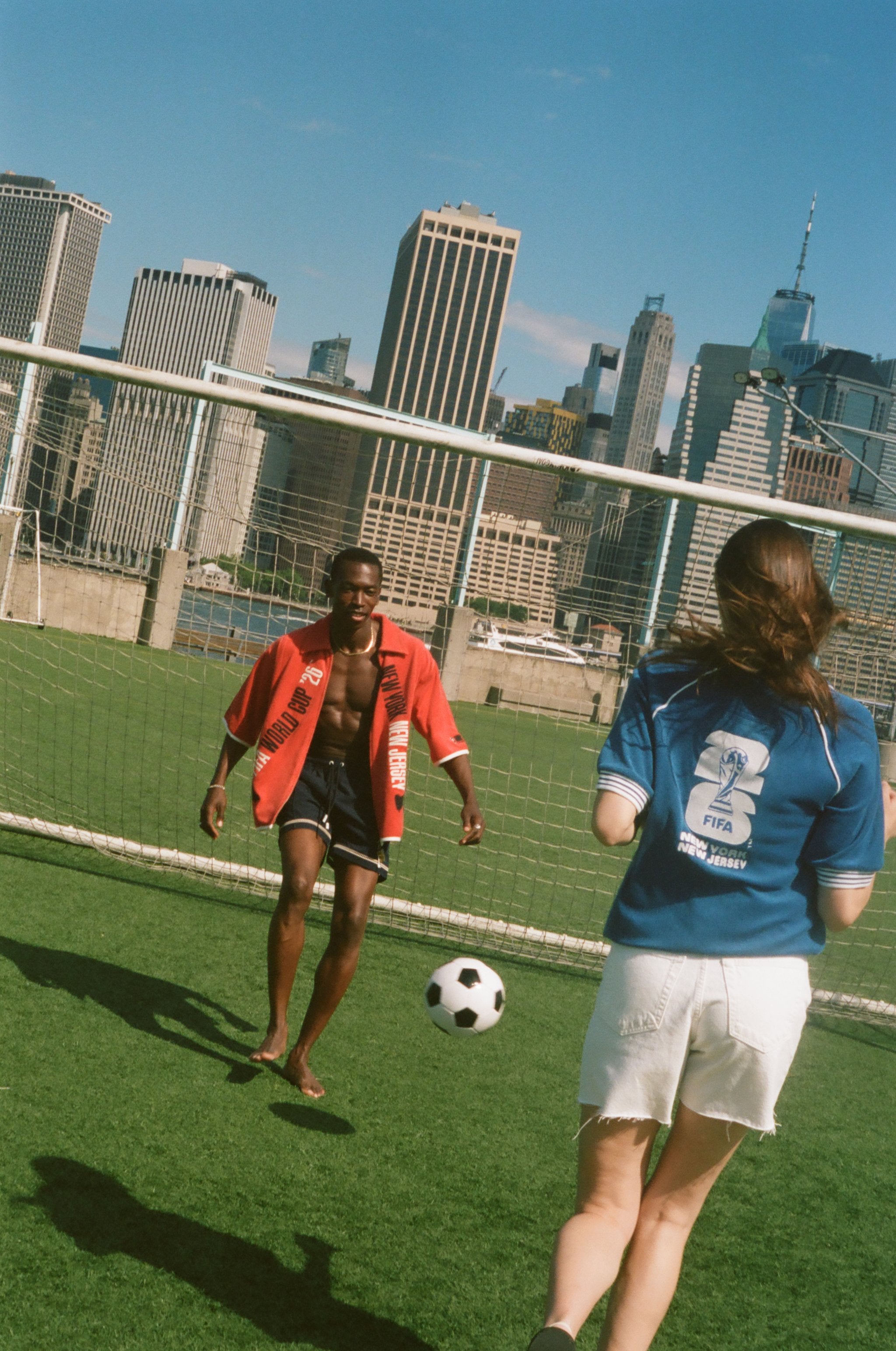 Two people playing soccer on a grassy field with a city skyline in the background, both wearing items from Tombolo and FIFA.