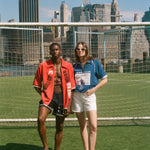 Two people standing on a soccer field with a city skyline in the background, wearing FIFA and Tombolo items. 