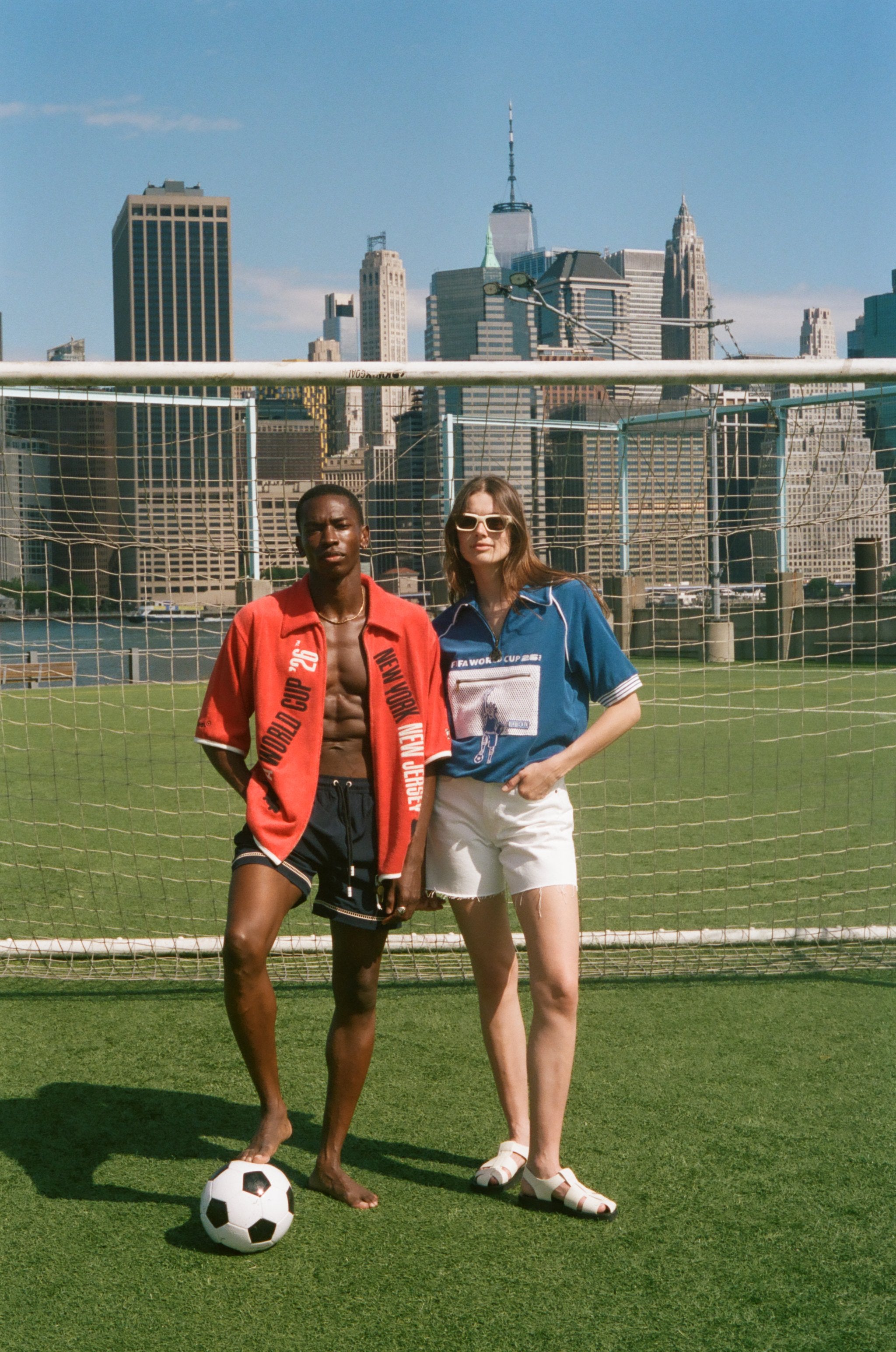 Two people standing on a soccer field with a city skyline in the background, wearing FIFA and Tombolo items. 