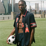 Man holding a soccer ball on a sports field with city skyline in the background