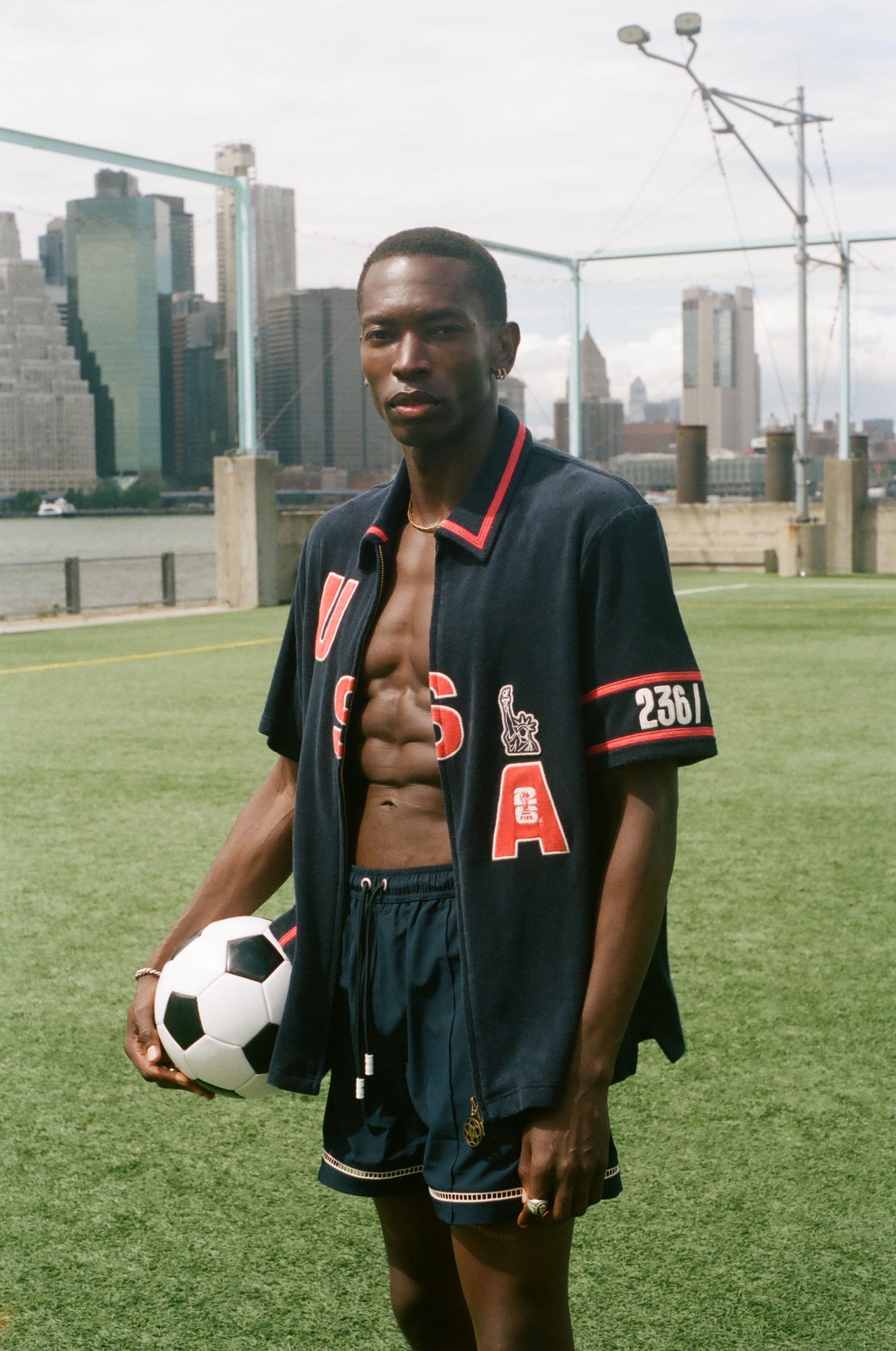 Man holding a soccer ball on a sports field with city skyline in the background