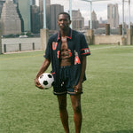 Man holding a soccer ball on a sports field with a city skyline in the background