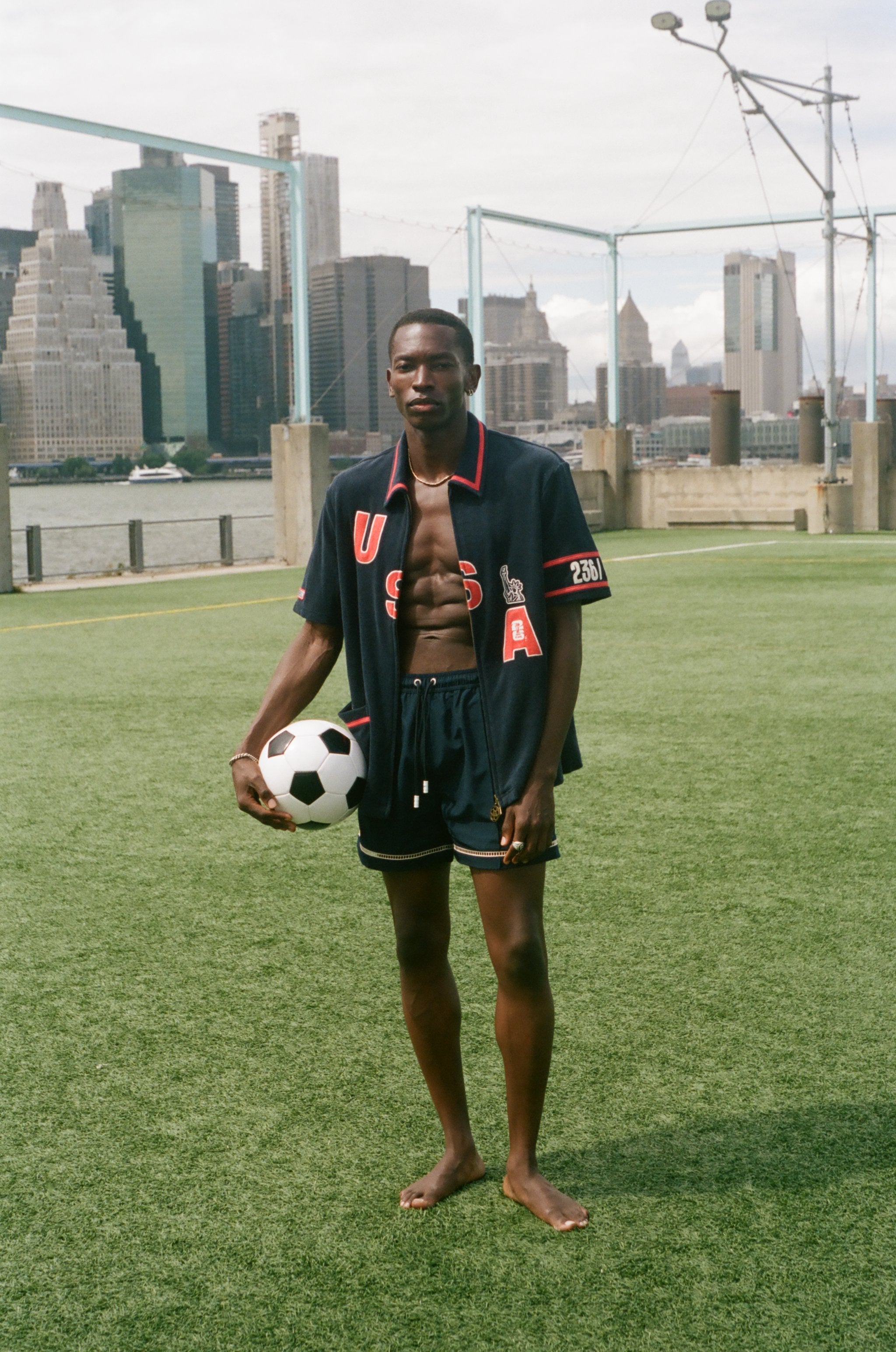 Man holding a soccer ball on a sports field with a city skyline in the background