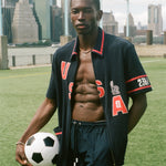 Man holding a soccer ball on a sports field with city skyline in the background, wearing the navy USA cabana. 