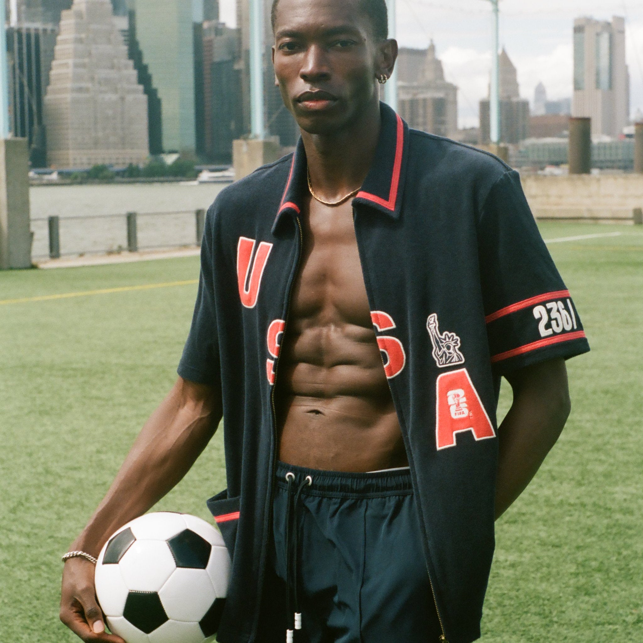 Man holding a soccer ball on a sports field with city skyline in the background, wearing the navy USA cabana. 