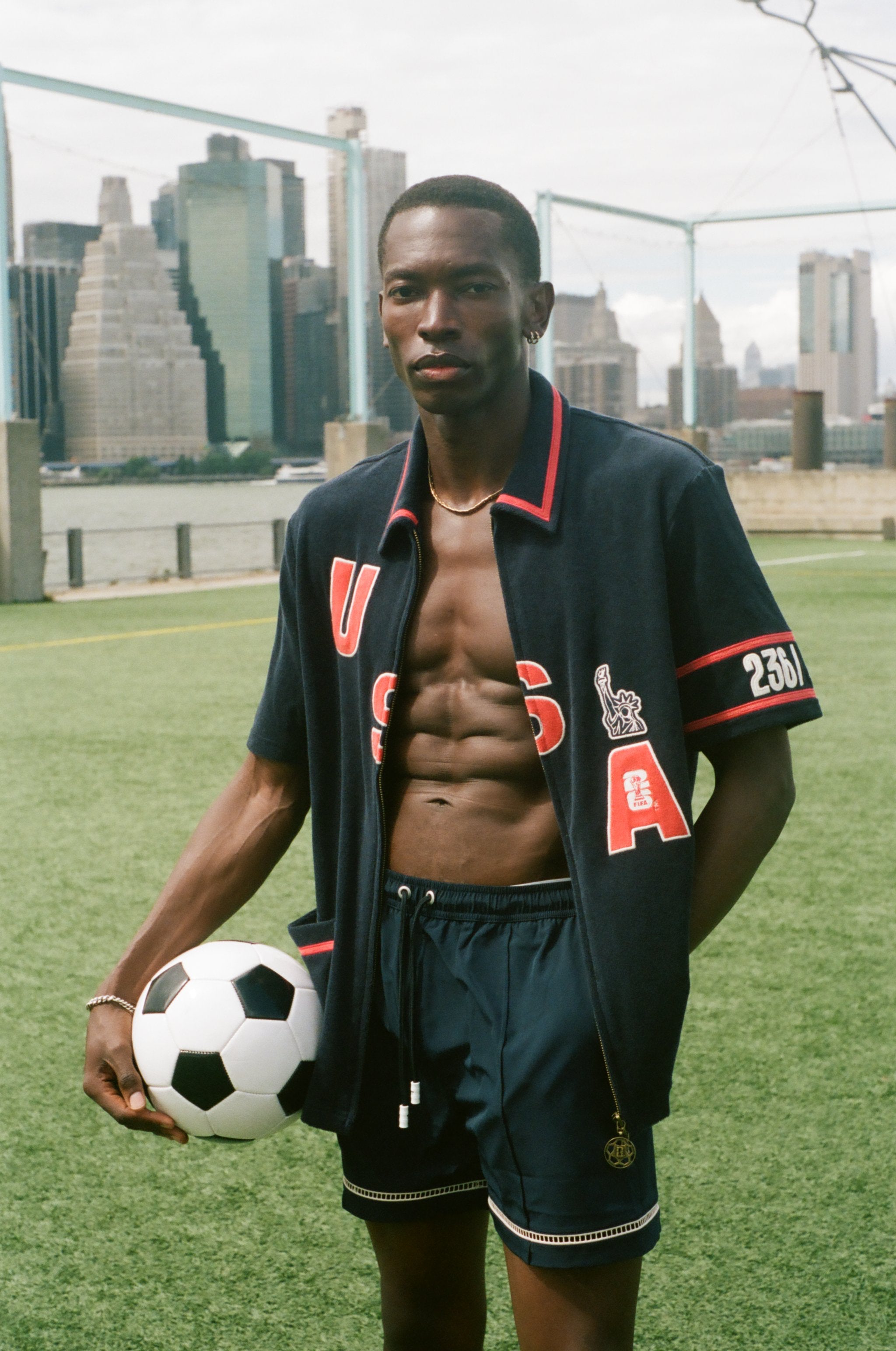 Man holding a soccer ball on a sports field with city skyline in the background, wearing the navy USA cabana. 