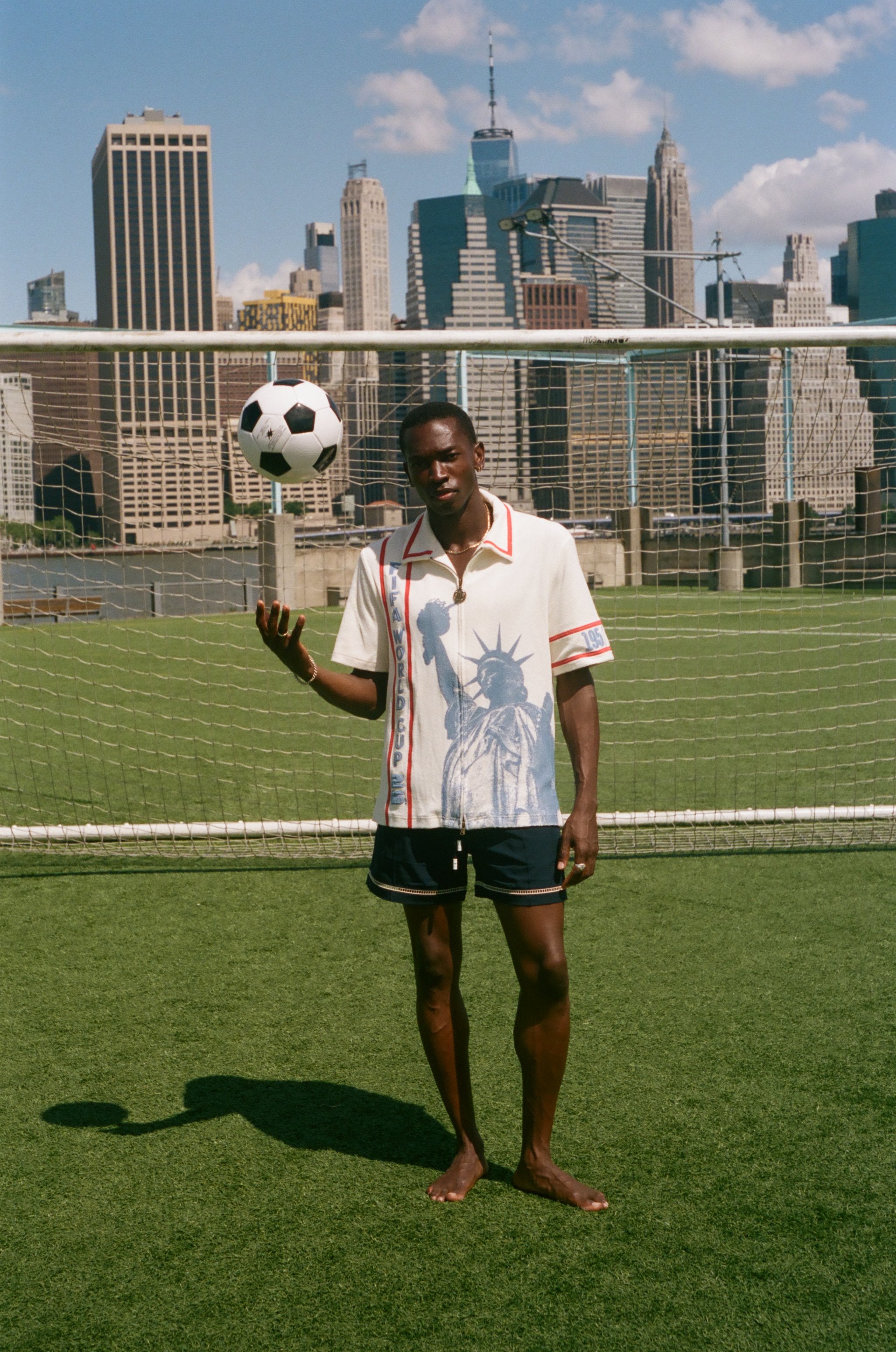 Model wearing a cream terry cloth zip-front shirt with red trim and a blue Statue of Liberty graphic, standing on a soccer field with a soccer ball and the New York City skyline in the background.