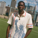 Model wearing a cream terry cloth zip-front shirt with red trim and a blue Statue of Liberty graphic, standing on a soccer field with a soccer ball and the New York City skyline in the background.