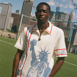 Model wearing a cream terry cloth zip-front shirt with red trim and a blue Statue of Liberty graphic, standing on a soccer field with a soccer ball and the New York City skyline in the background.
