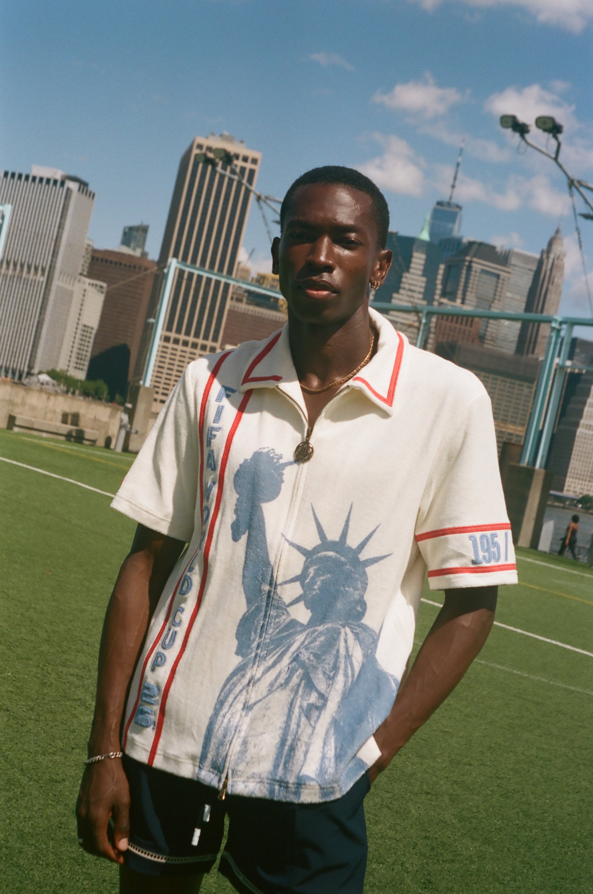 Model wearing a cream terry cloth zip-front shirt with red trim and a blue Statue of Liberty graphic, standing on a soccer field with a soccer ball and the New York City skyline in the background.