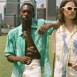 Two people standing in a grassy area with a city skyline in the background, wearing items from the Tombolo and FIFA collection.