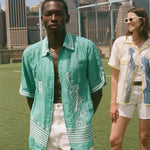 Two people standing on a soccer field, wearing the Teal Stars and Stripes Cabana and the Lady Liberty Hawaiian shirt from the FIFA and Tombolo collection.