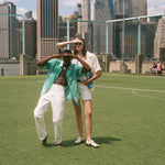Man and woman standing on a soccer field, looking off into the distance. Both are wearing items from the FIFA and Tombolo collection.
