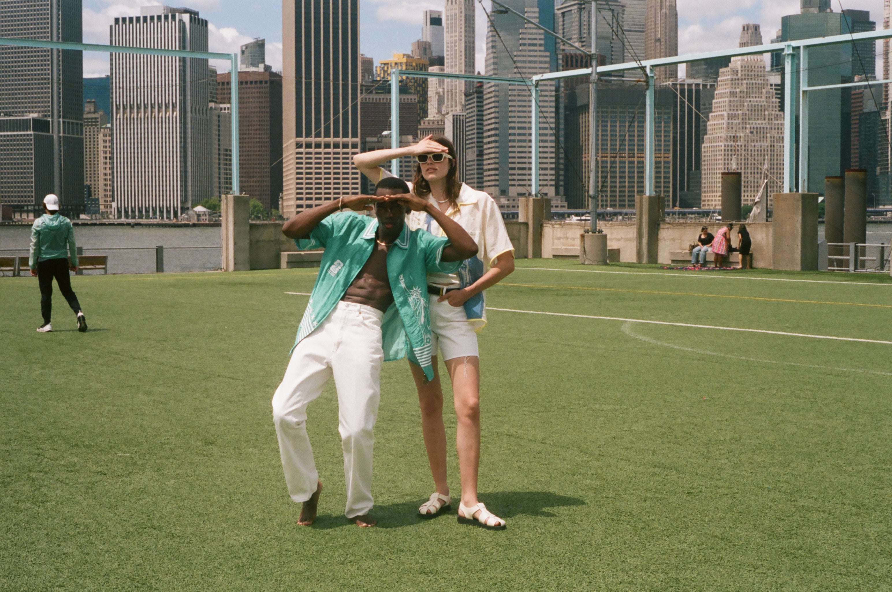 Man and woman standing on a soccer field, looking off into the distance. Both are wearing items from the FIFA and Tombolo collection.