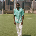 Man wearing the teal stars and stripes cabana with white pants, standing on a soccer field with a city skyline in the background. 